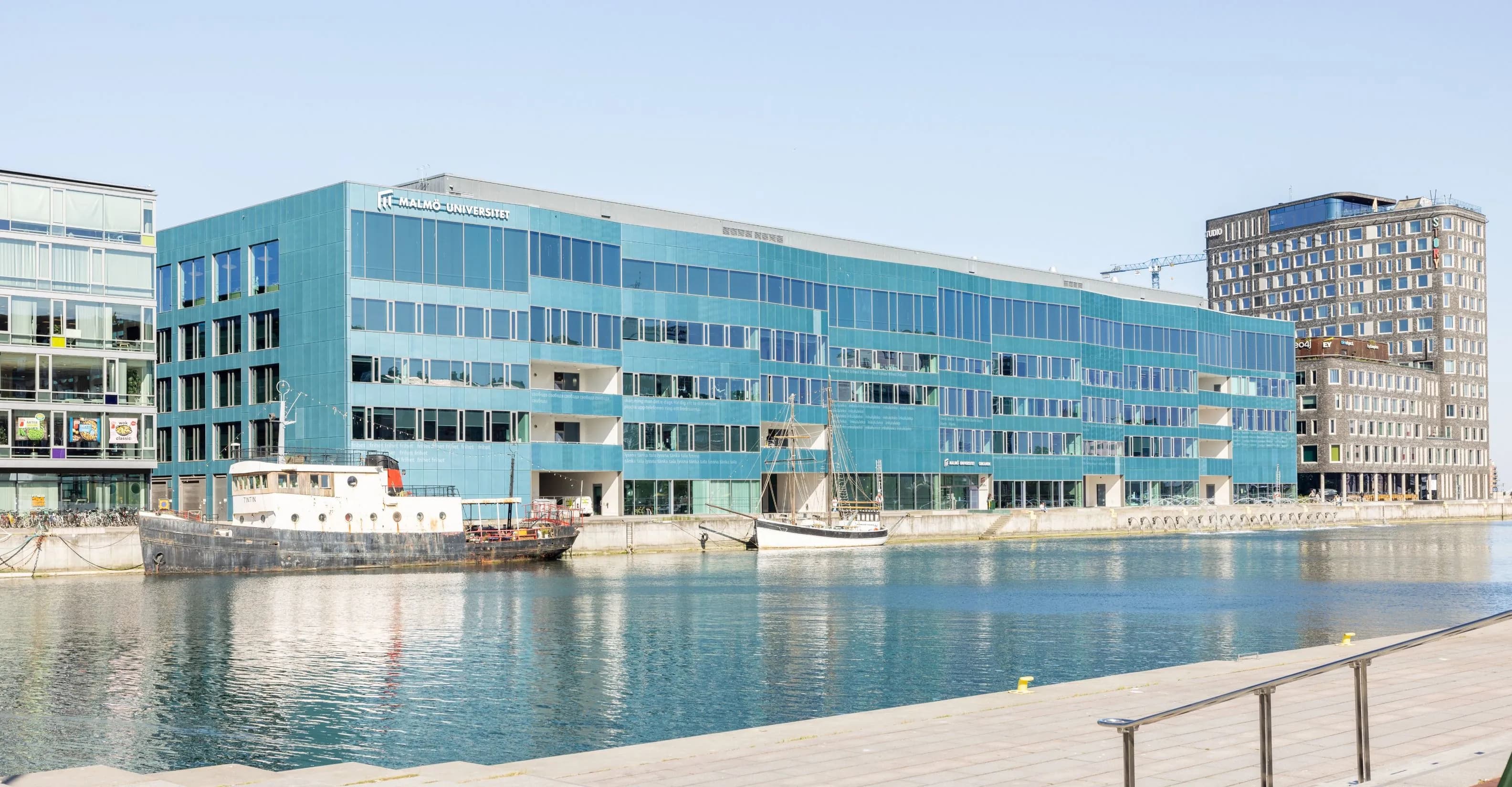 Modern waterfront building of Malmö University with a turquoise glass facade, located along a canal with docked boats, under a clear blue sky.
